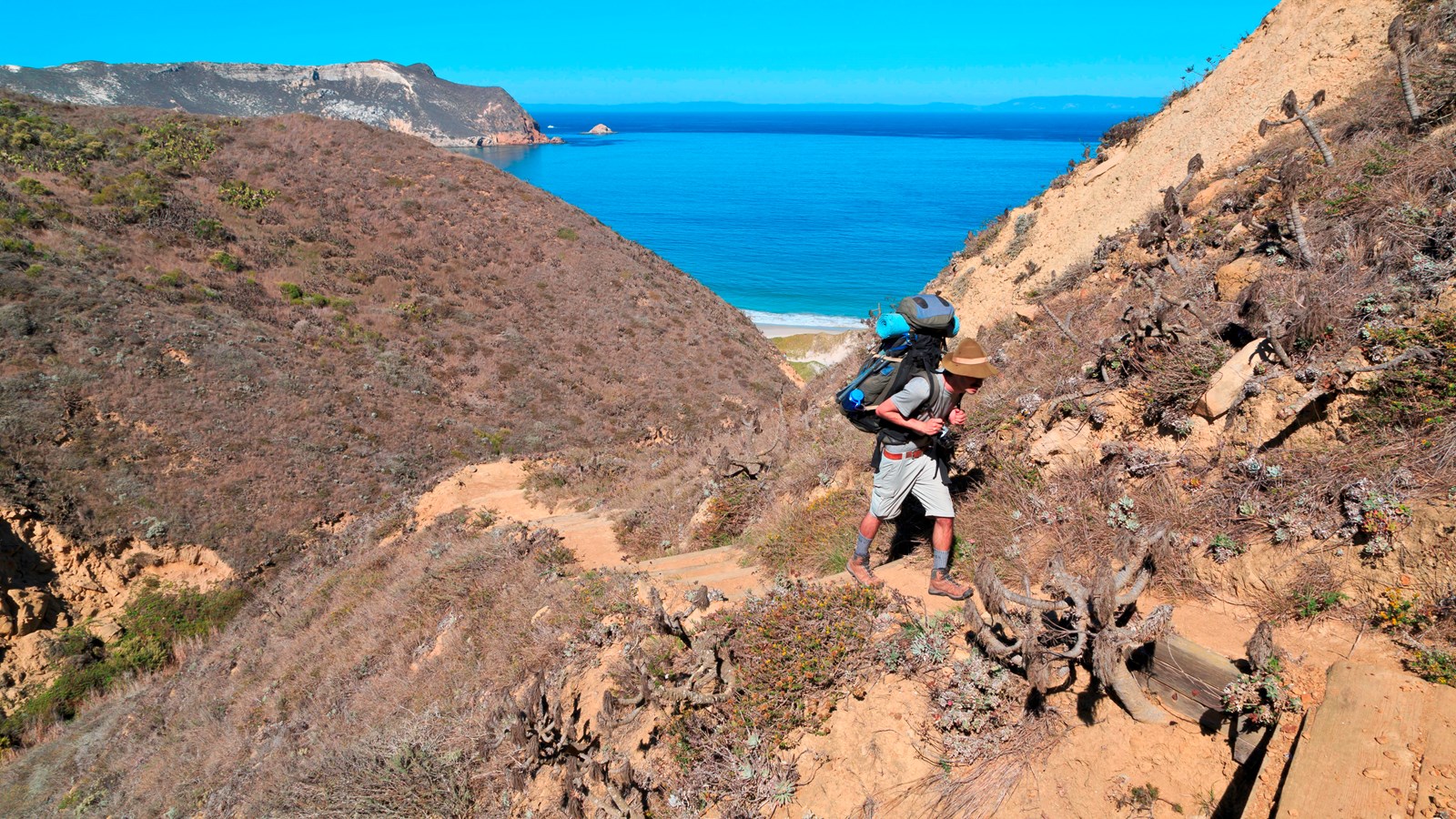 Man hiking with backpack up steep trail from beach. 