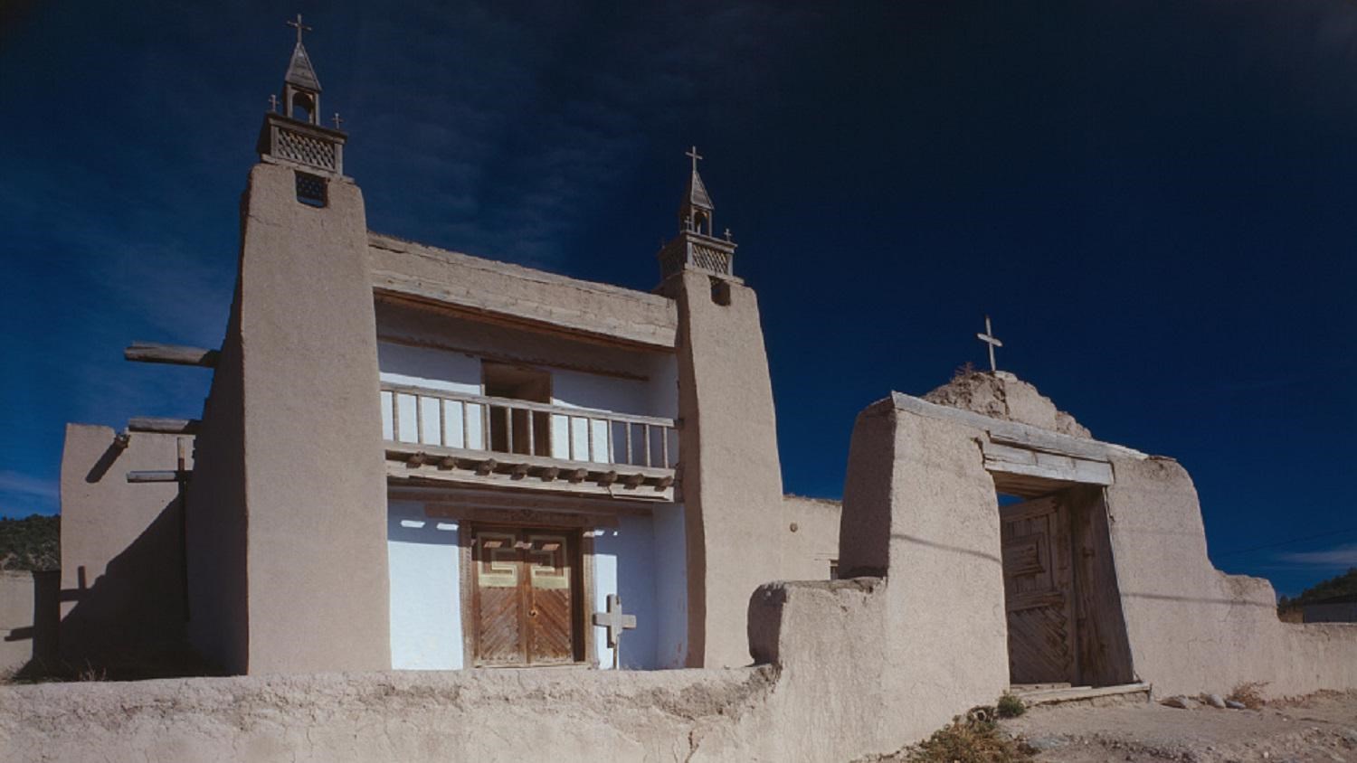 Two story adobe building with tan towers on either side of a white washed  door and balcony.