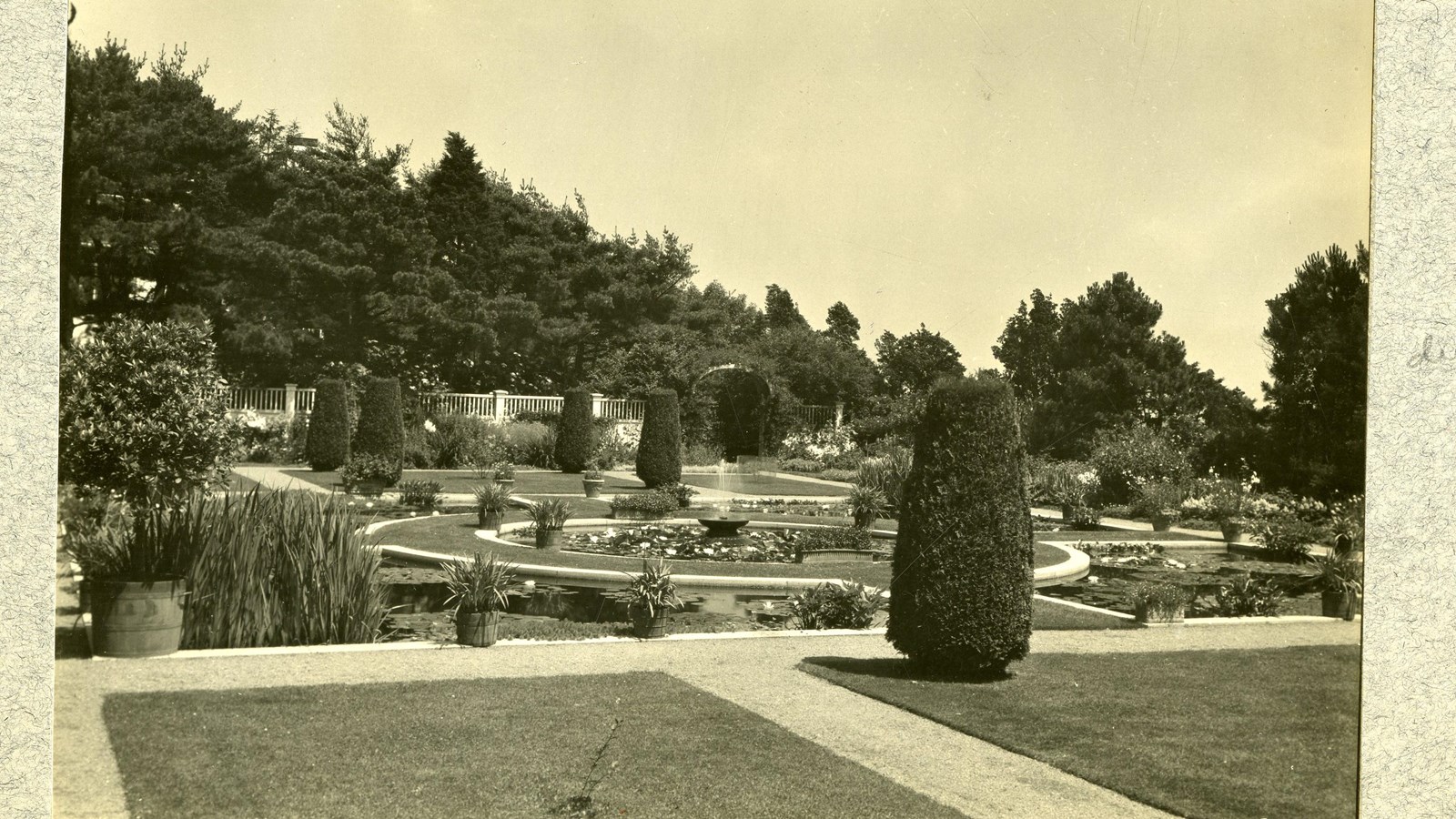 Black and white of flat grassy area with plants and flowers along paths, fountain in middle