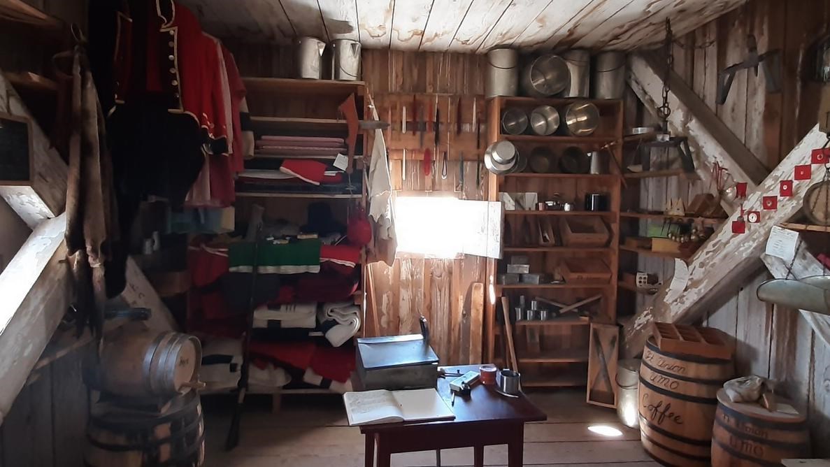 Wooden walls with shelves containing tin, metal and fabric items. Wooden barrels. Desk with inkpot. 