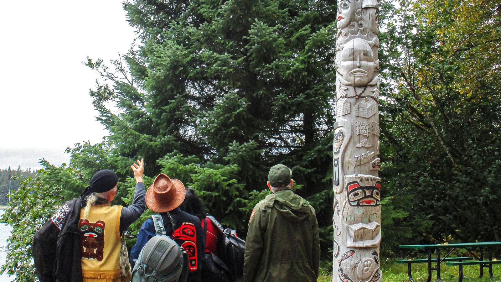 Several people with backs to the camera look at the healing totem pole while one points at it.