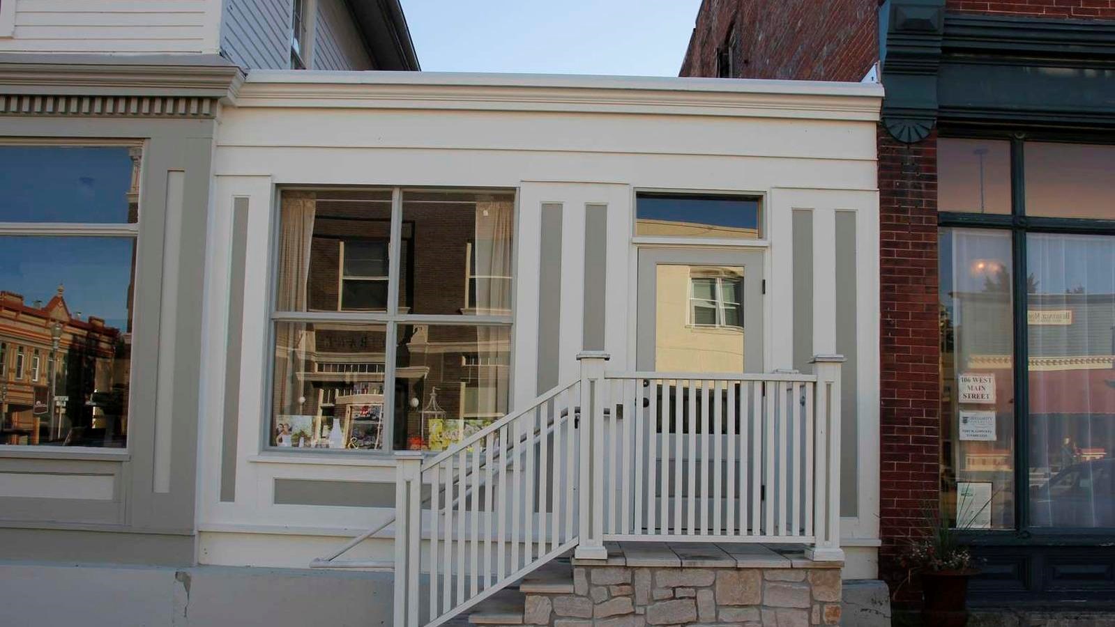 A white one-story building has a storefront window and larger buildings flush on each side.