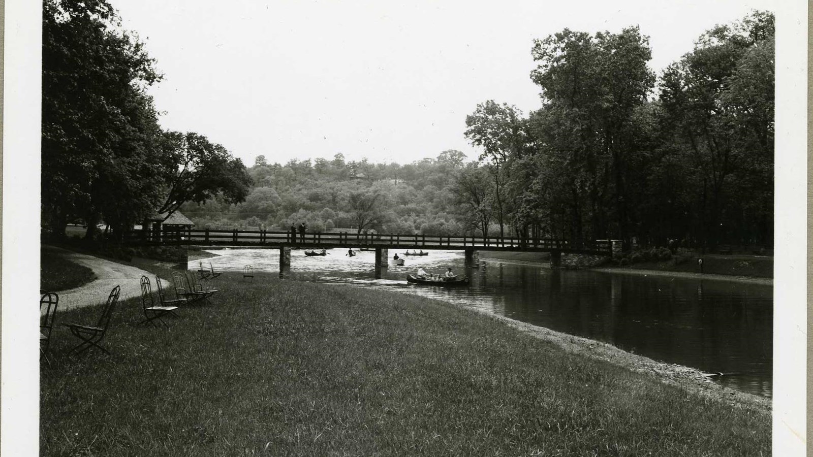 Black and white of flat grassy area next to body of water with bridge over, people boating on water