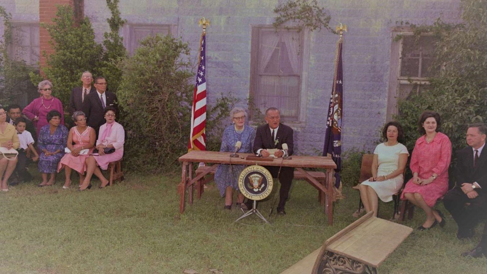 In front of a grey building, the president makes remarks to a seated group.