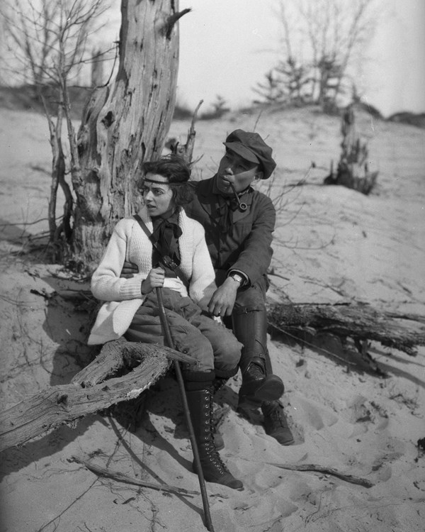 Historic black and white photograph of man & woman in hiking gear taking a break on a log on a dune.
