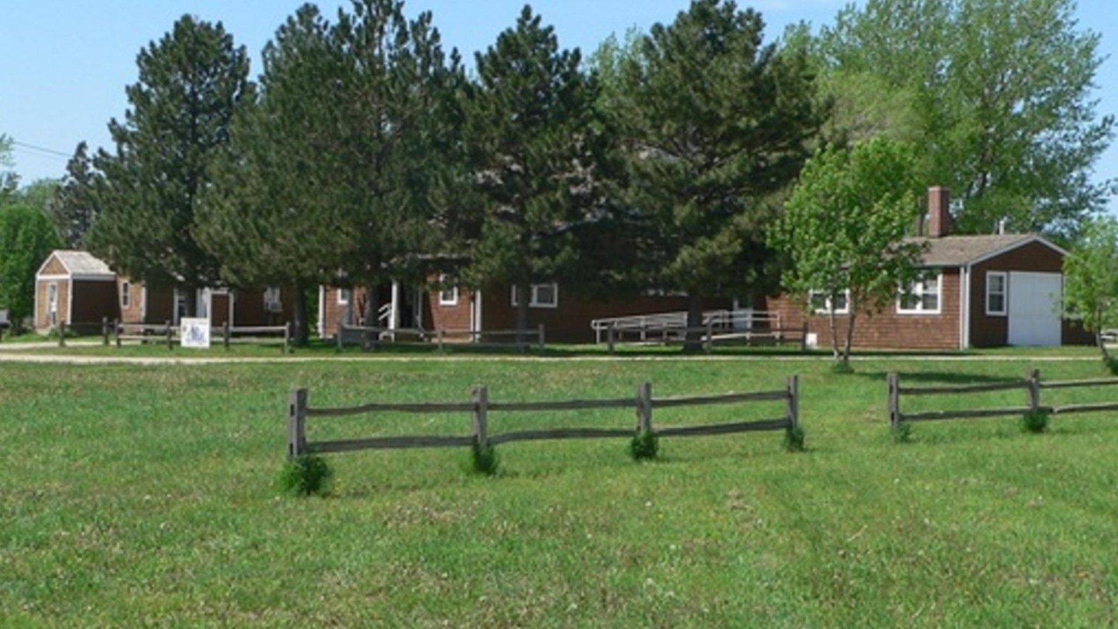 A split rail fence stands on a green lawn in front of a red building with white trim