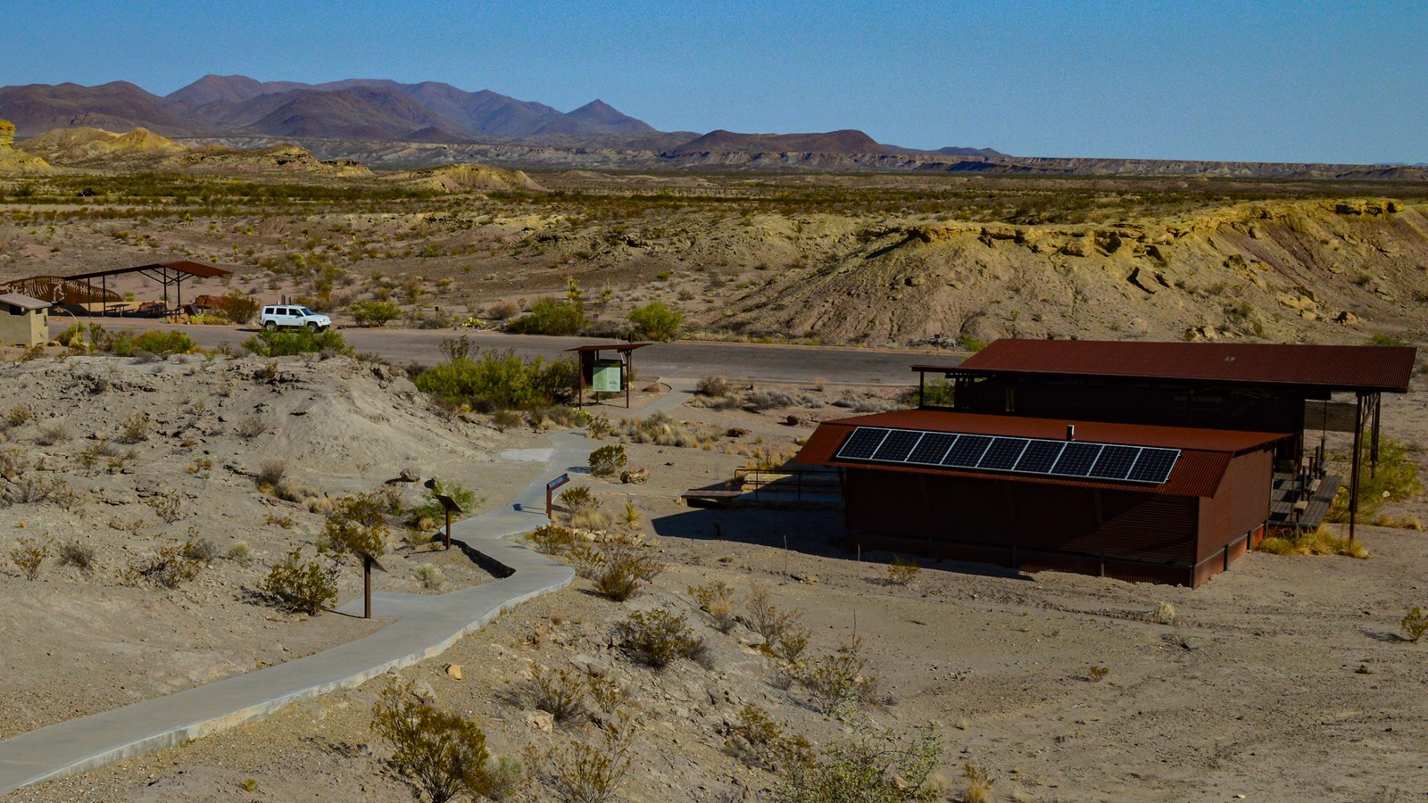 A metal building lies next to a paved trail that climbs a hill to several information exhibits.