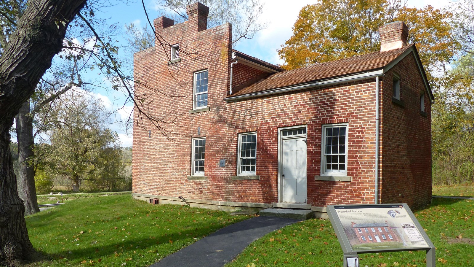 Paved path leads past a graphic panel to the white side door of an L-shaped brick house. Four window