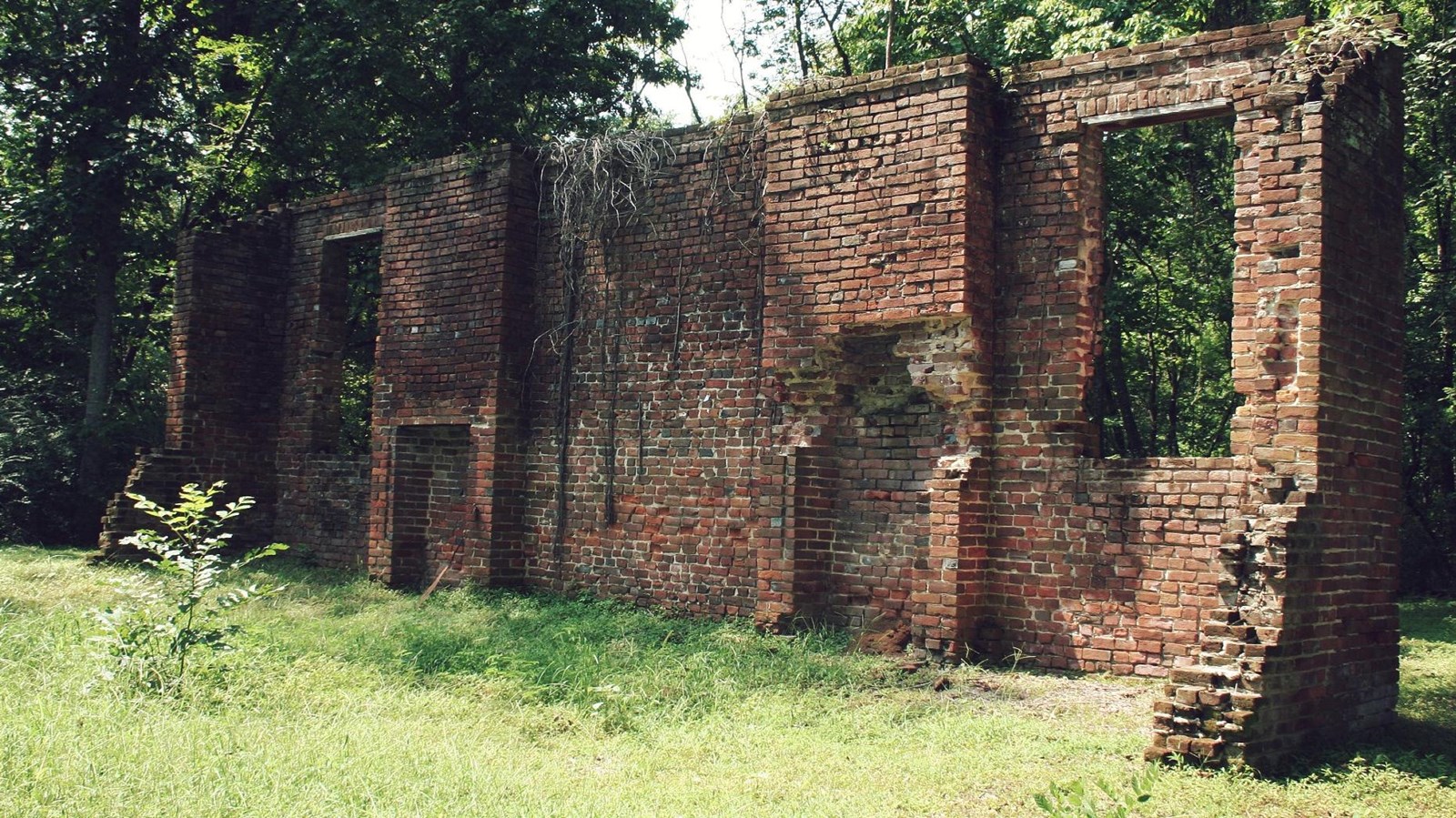 The ruins, just a brick wall, stands amongst green trees. 
