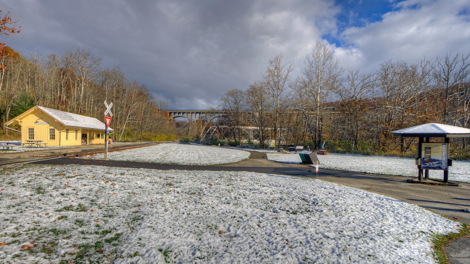 Yellow train station & tracks to left of distant arched bridge in snow, three-sided kiosk at right.