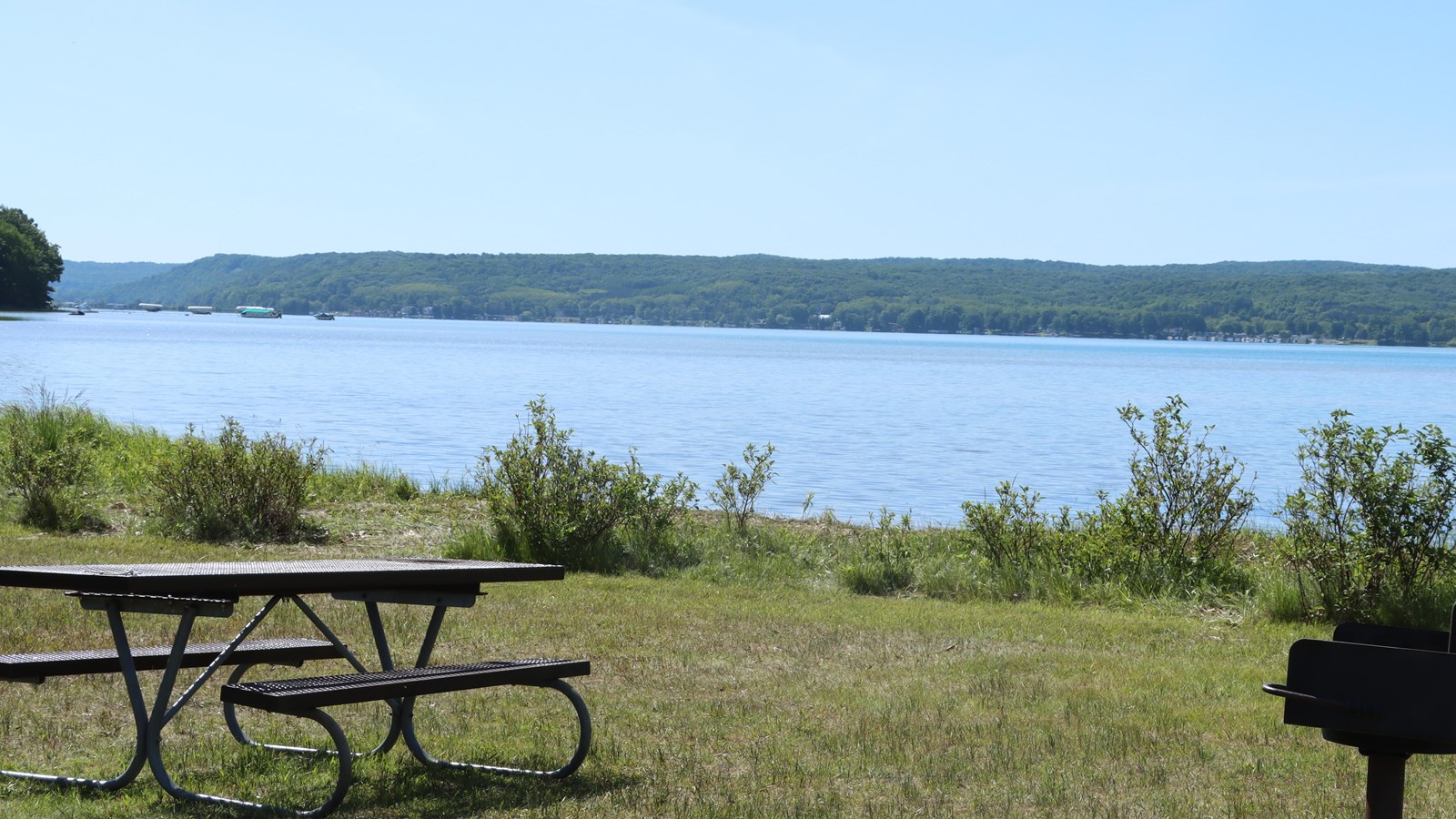 The clear blue lake opens from a grassy shoreline with a few small shrubs, picnic table and grill.