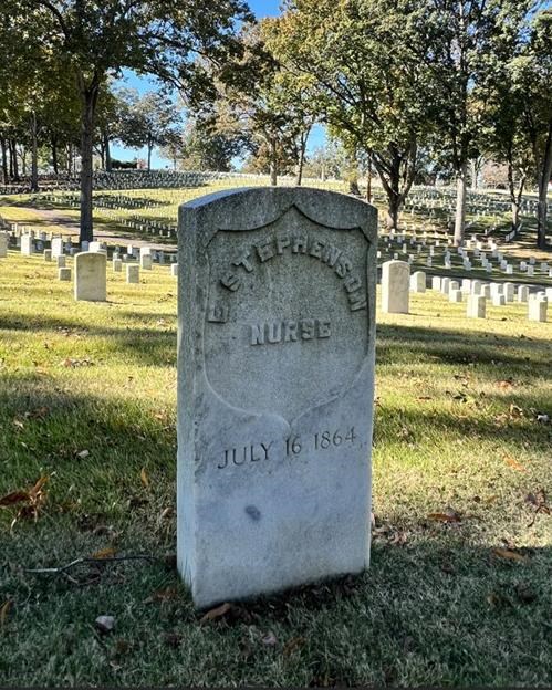 White headstone with trees, blue sky, and other white headstones in background.