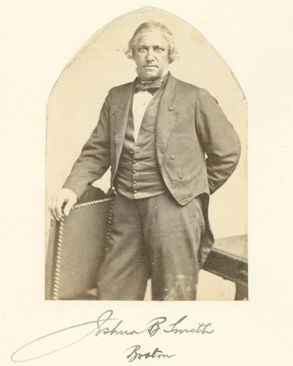 Historical photo of a man in a suit standing next to a table with his right arm resting on it.