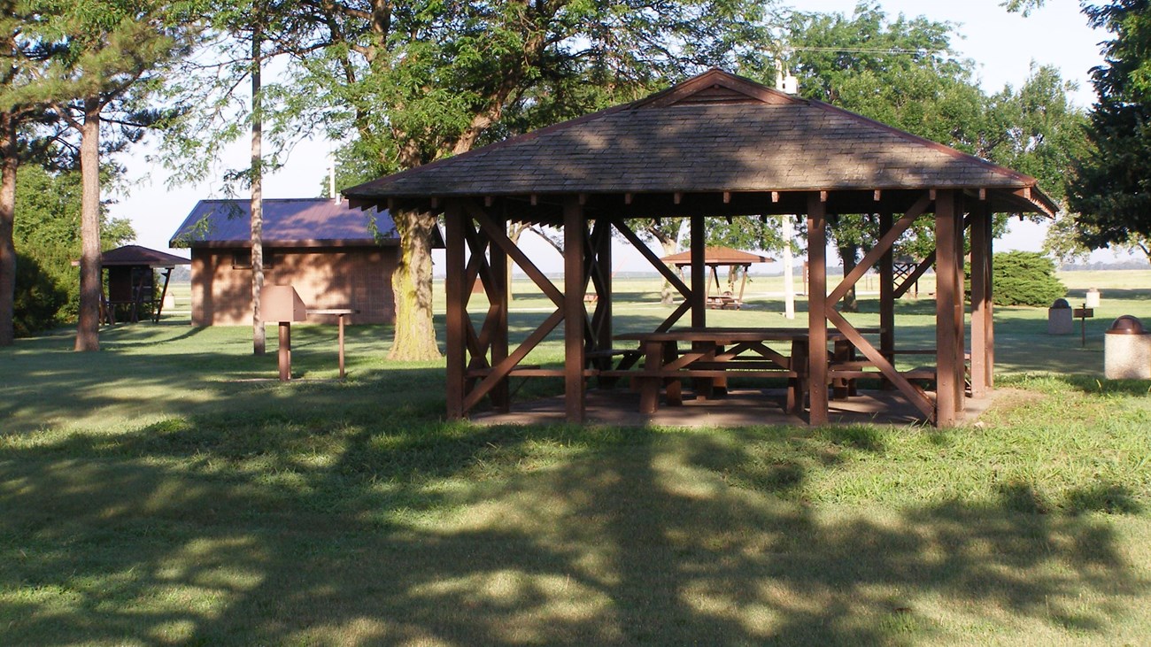 Picnic table under a shelter surrounded by trees with brick restroom in background.