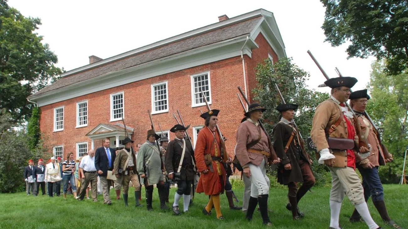 A line of people dressed as 18th C militia and in modern clothes march past a large brick house.