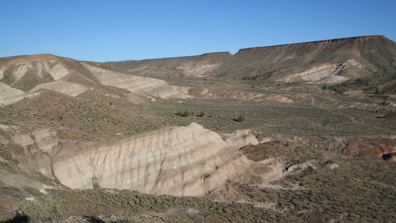 A tan geologic formation sits in a valley with plateaus in the background