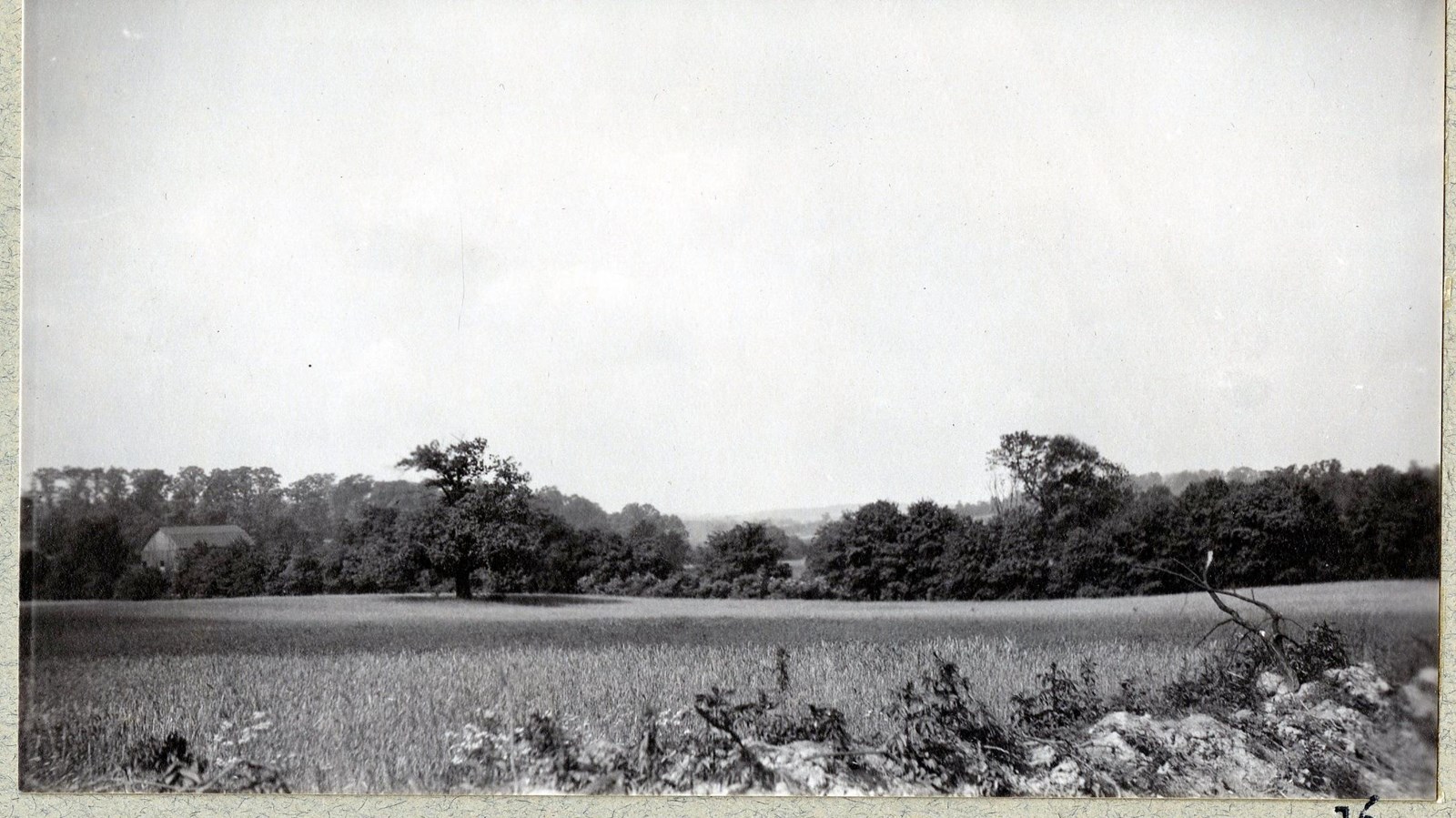 Black and white of flat grassy area with line of trees on the edges with a building in the trees 