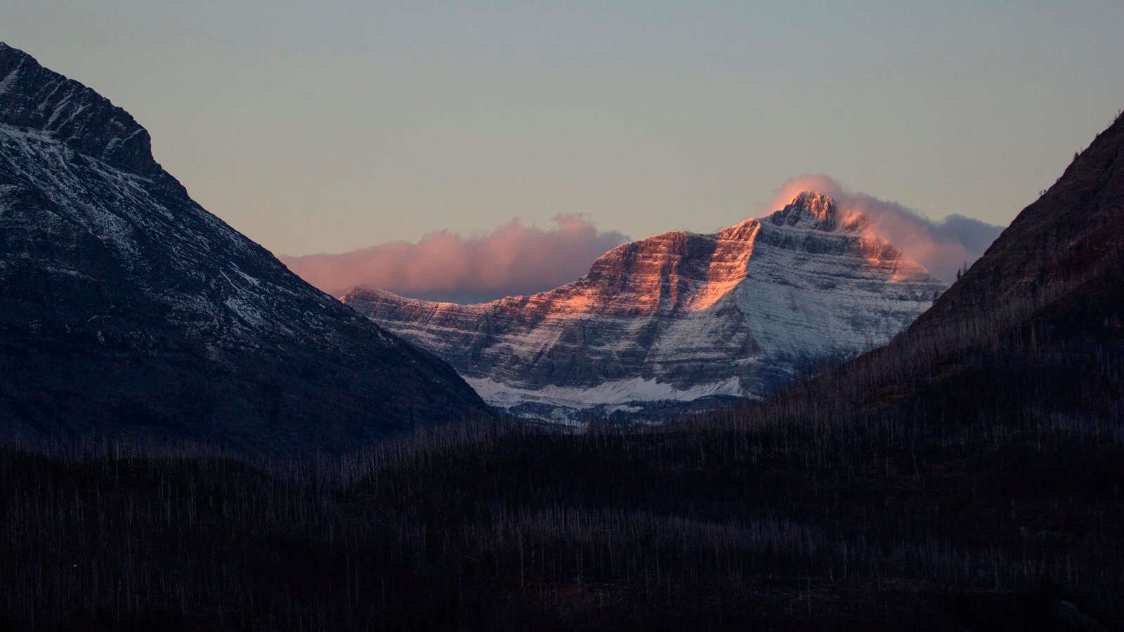 Two peaks of a mountain are colored pink by the sunset. A thin cloud shrouds the ridge in between.