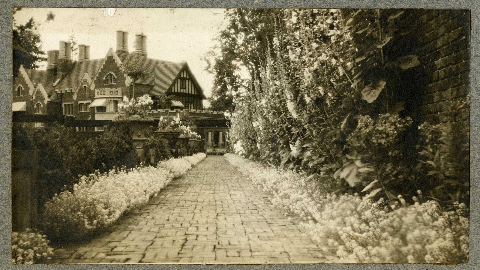 Black and white of brick path leading to large home lined with flowers and walls with plants