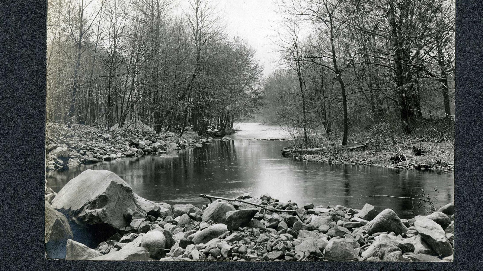 Black and white of river lined with rocks and trees. 