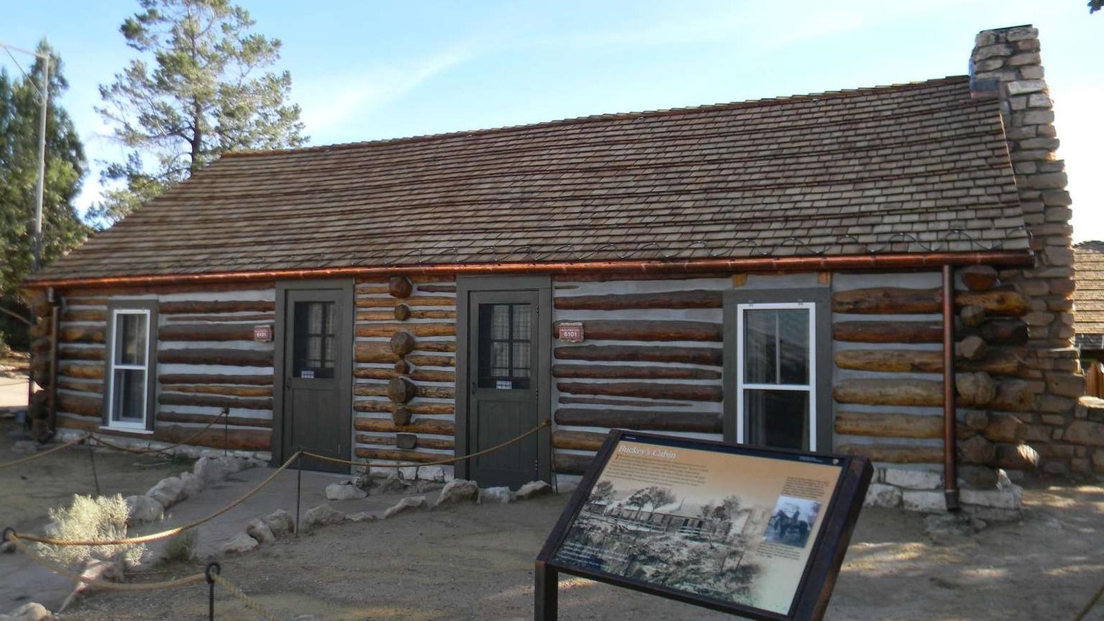 A dark brown log cabin with a stone chimney. A sign in front tells the story of its original owner.