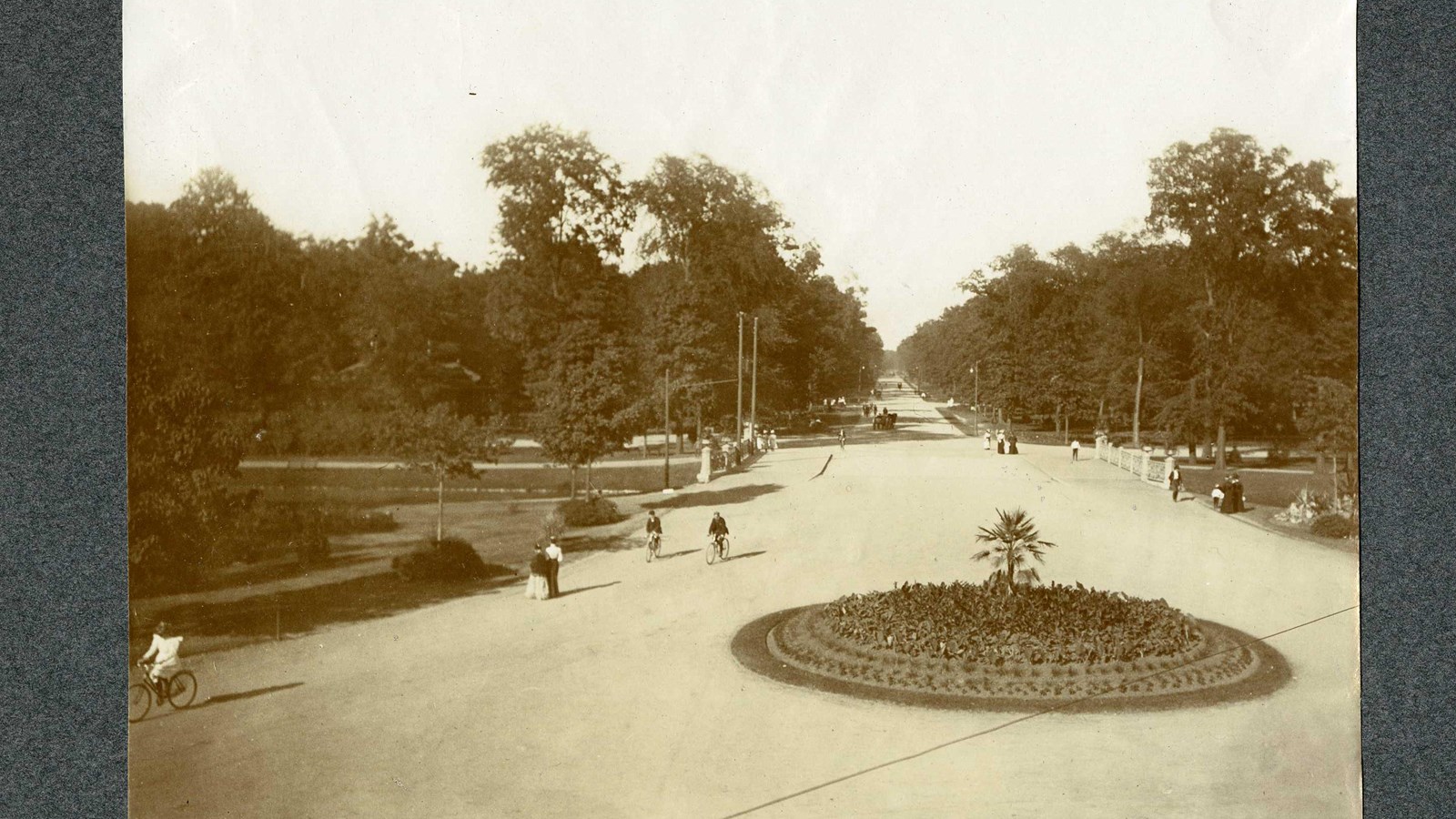 Black and white of dirt path lined with trees with circular plant area and people along the path. 