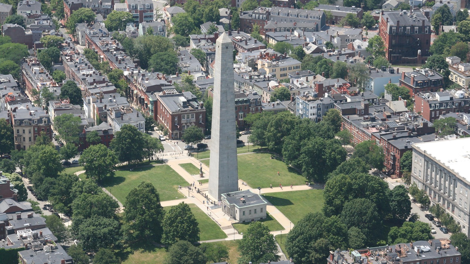 View of a dense neighborhood of mostly 3 story dwellings with green space and monument in center.