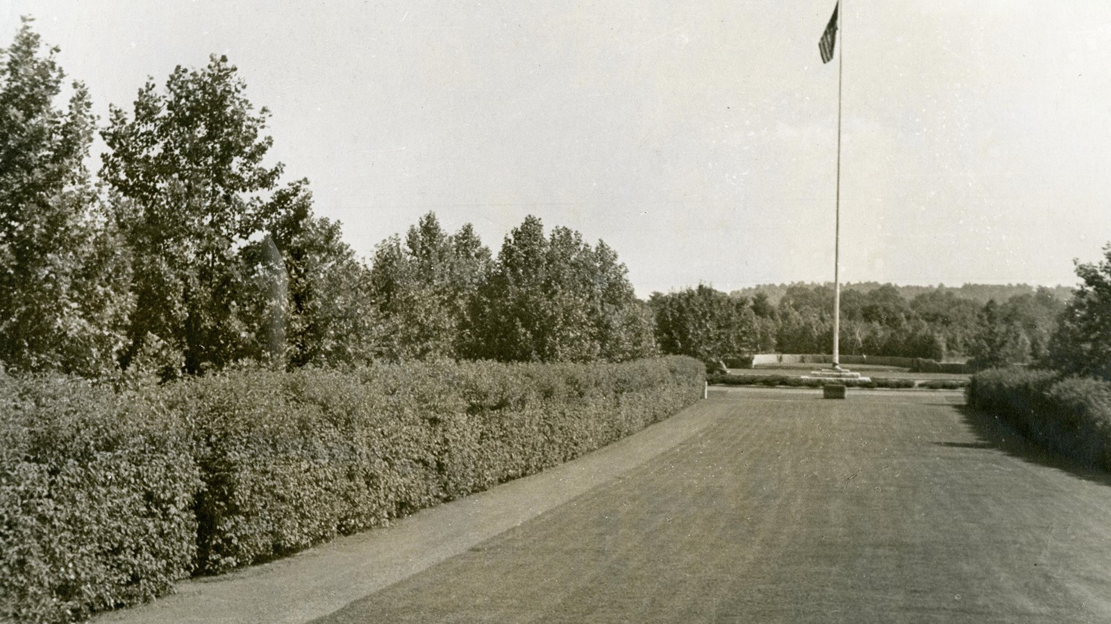 Black and white of flat grassy area lined with shrubs with pole and American flag at end