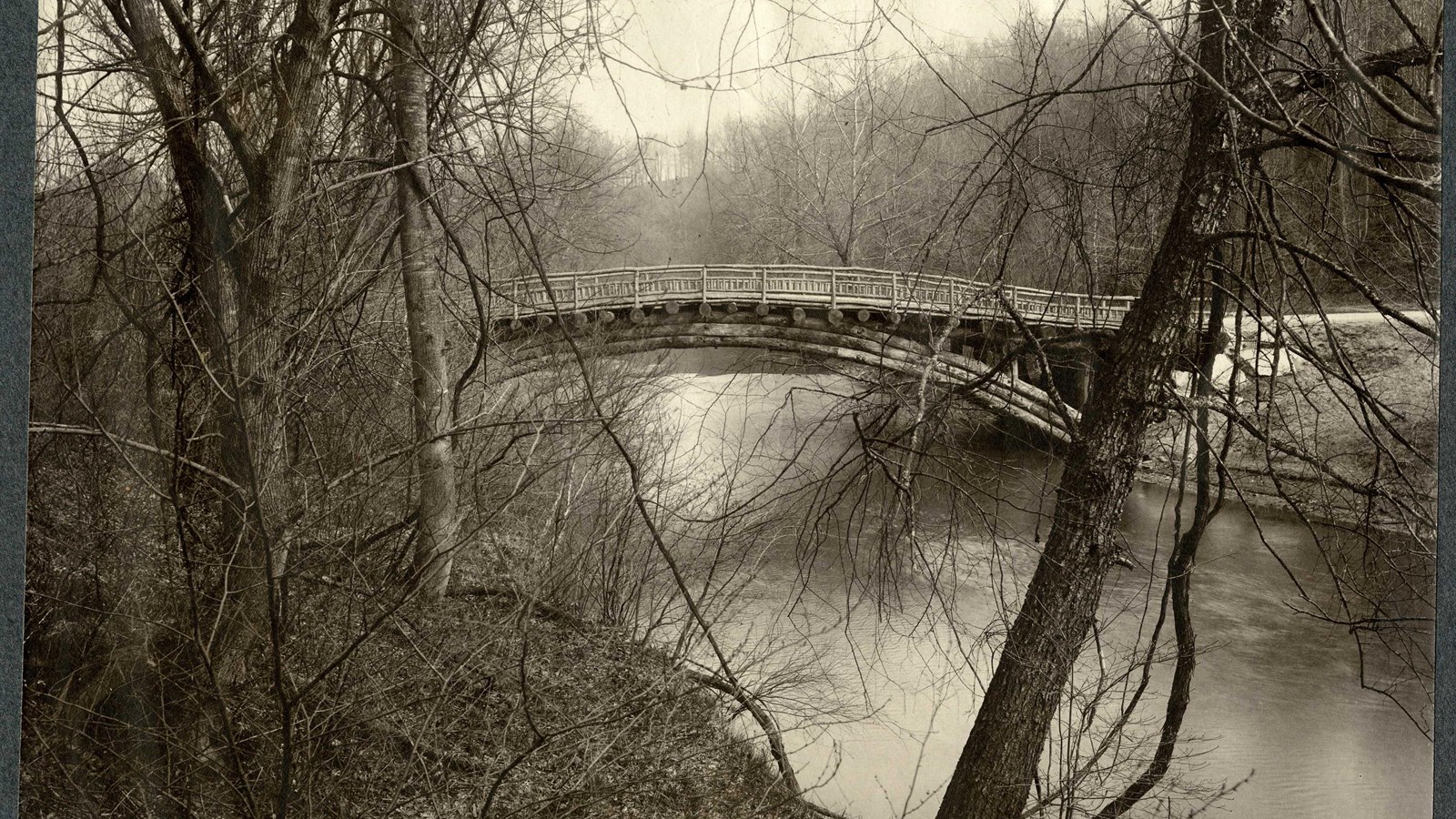 Black and white of bridge over water with trees on one side, hilly going down to water on other