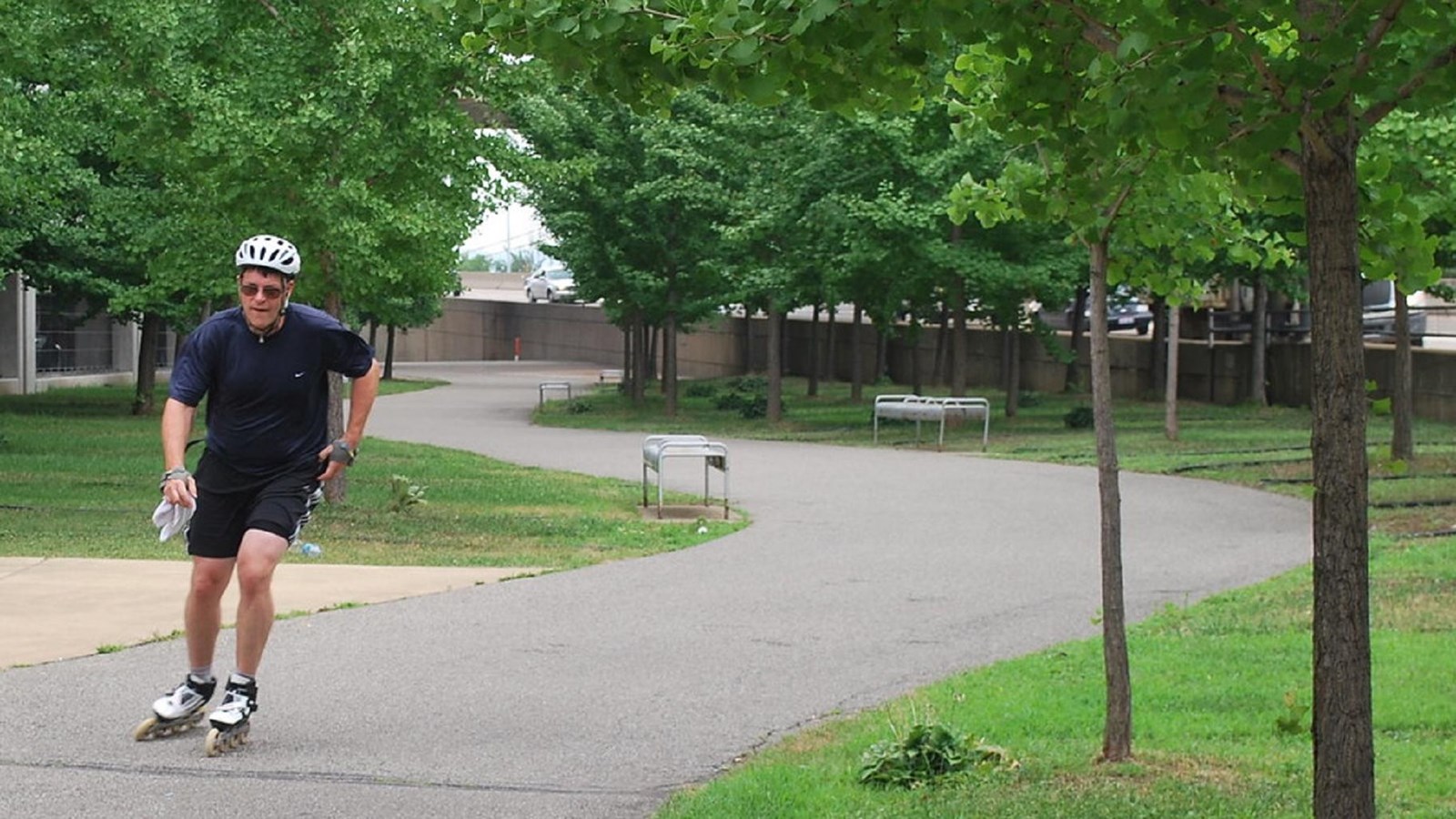 A man rollerblading on a paved trail through a tree-dotted city park