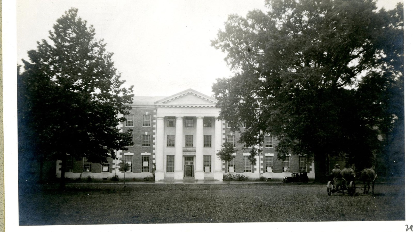 Black and white of large building on flat grassy area with trees on the edge of the building