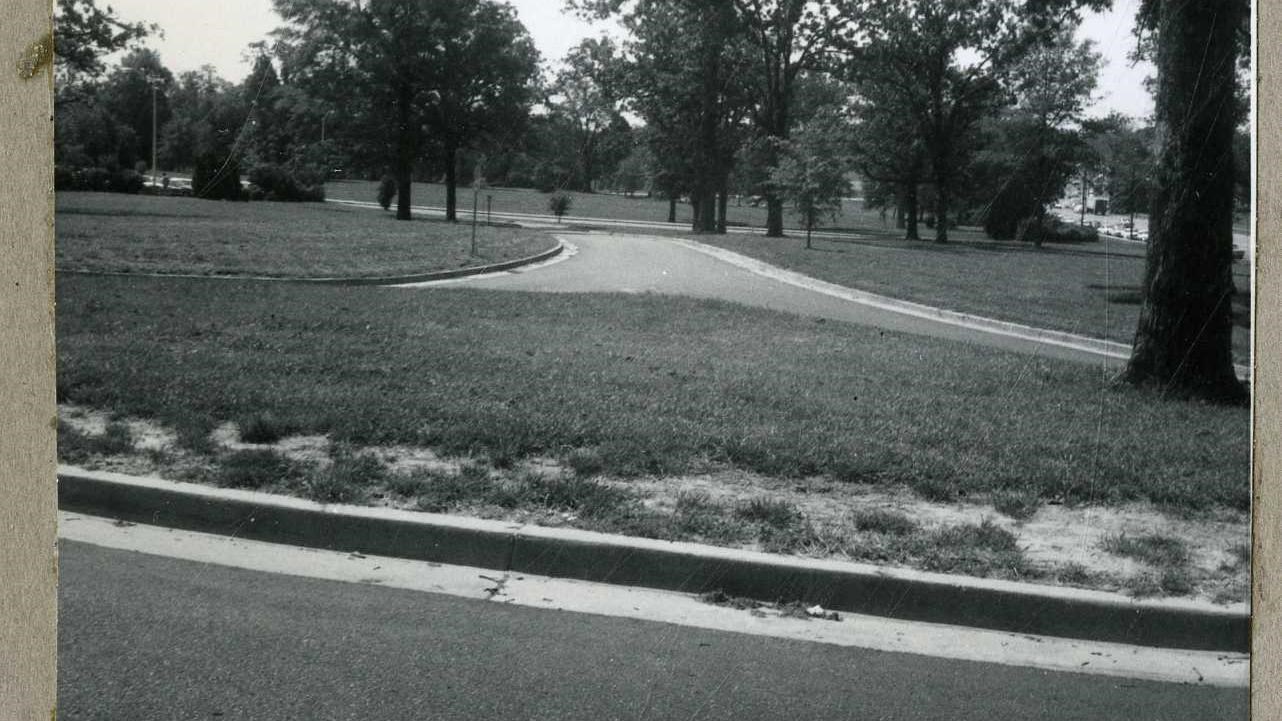 Black and white of road cutting through flat grassy area with some trees along edge of grass