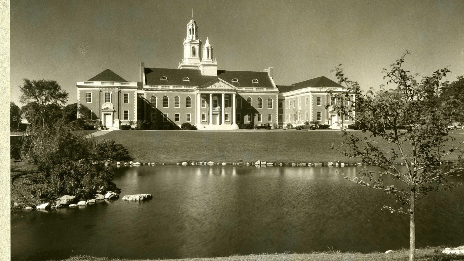 Black and white of body of water in front of grassy hill leading to a large building