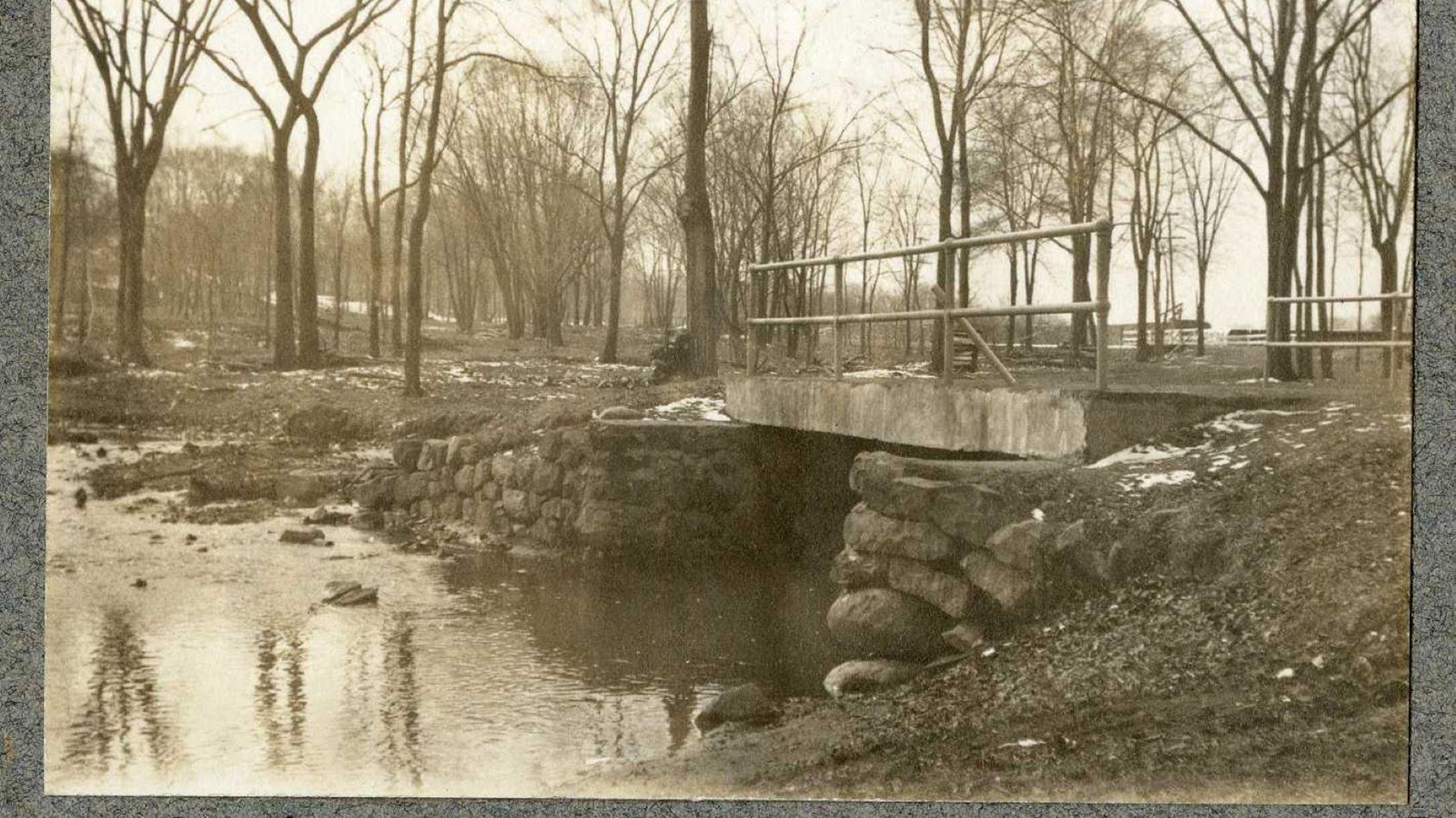 Black and white of small stone bridge over water with trees with no leaves on the other side 