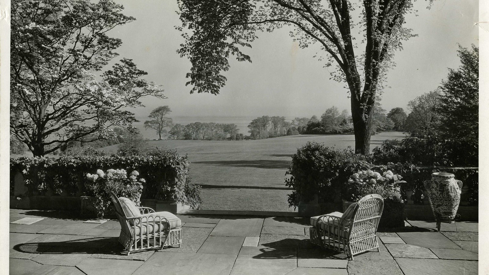 Black and white of two chairs on brick patio facing large flat open grassy area, trees on edge