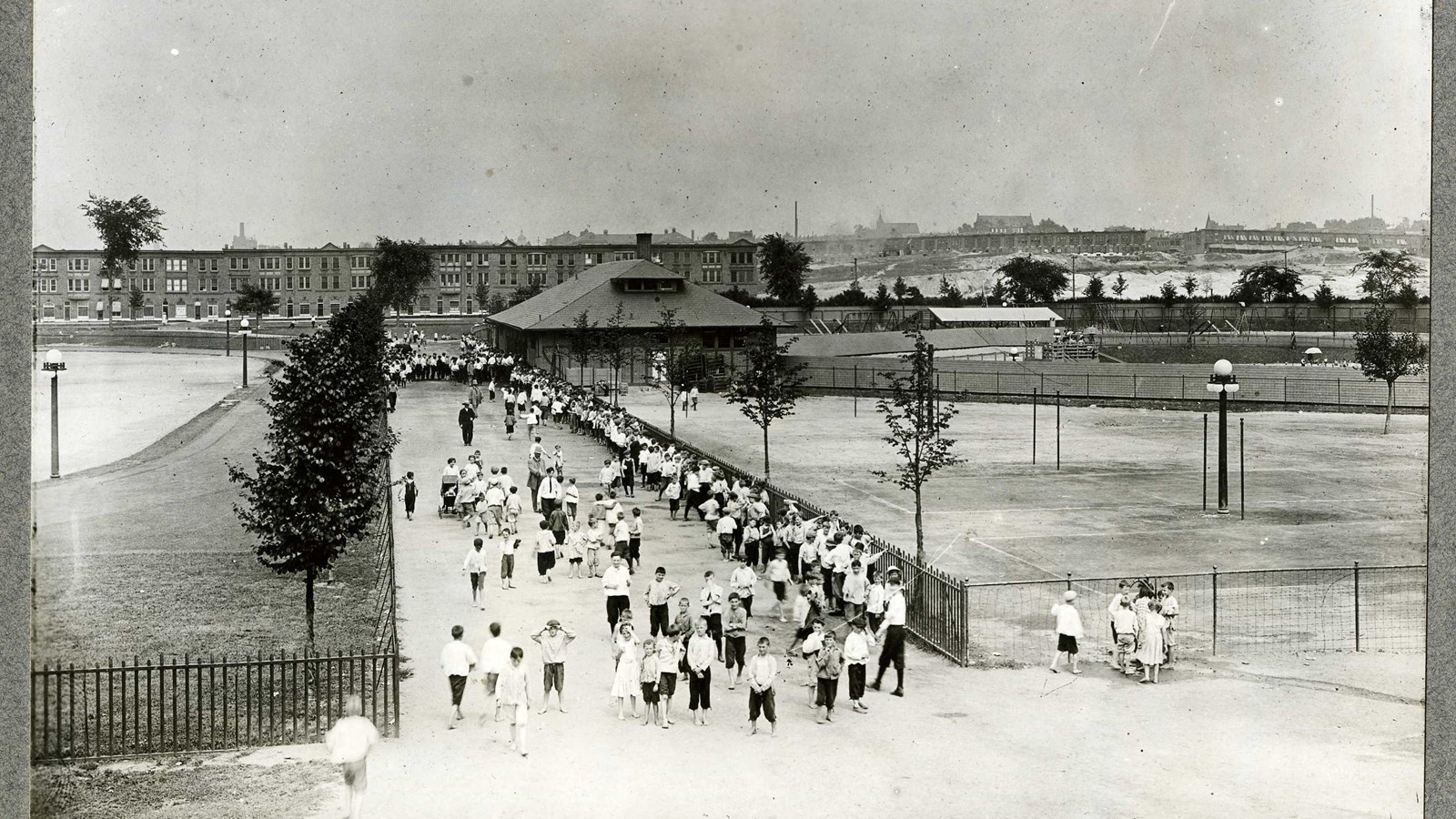 Black and white of dirt path with many people on it, with city in the background 