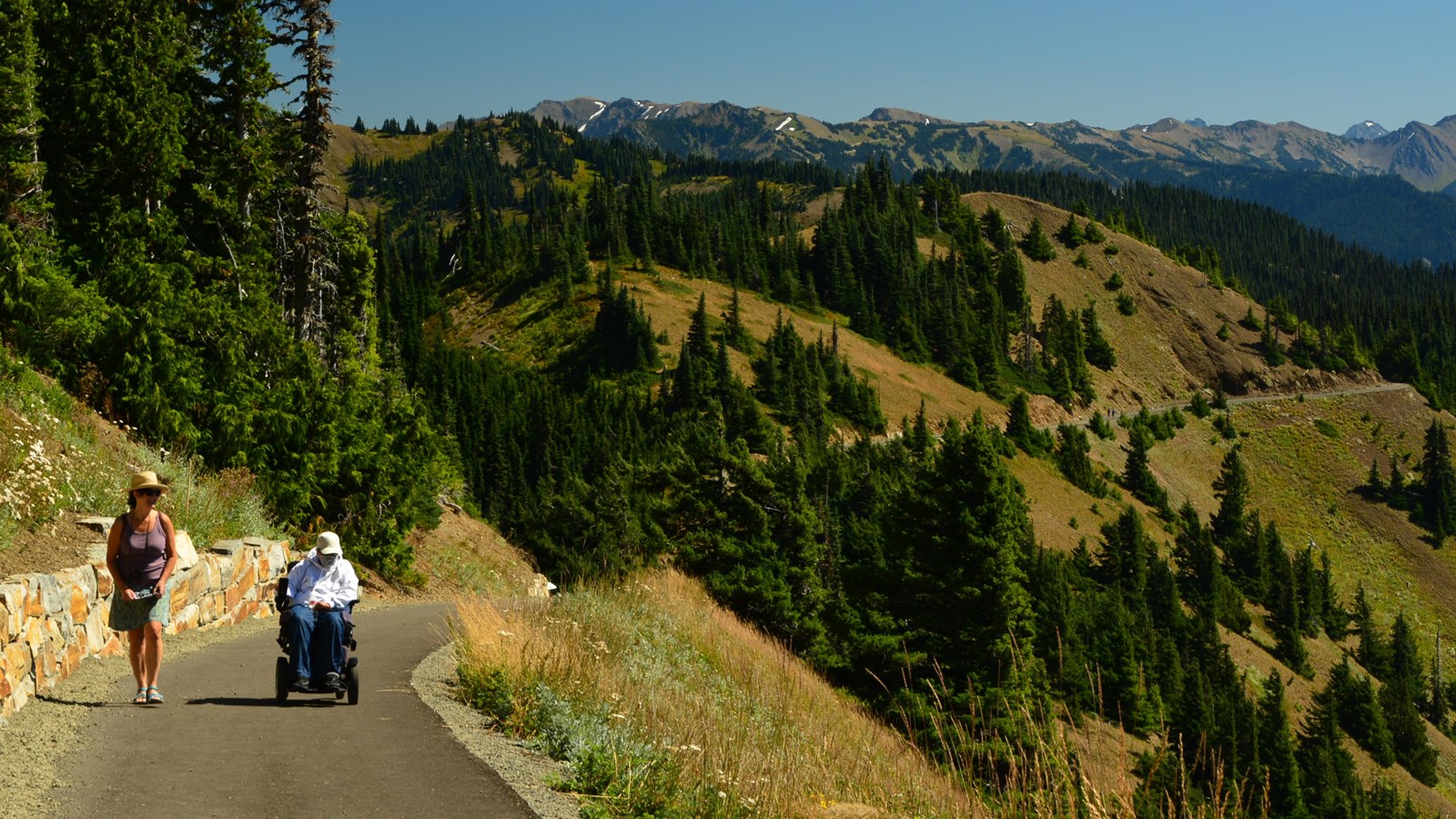 A hiker and wheelchair user on a paved trail with mountains in the background.
