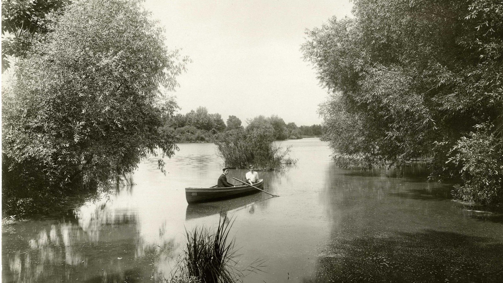 Black and white of two people in canoe on water with trees leaning over side, some grass in water 