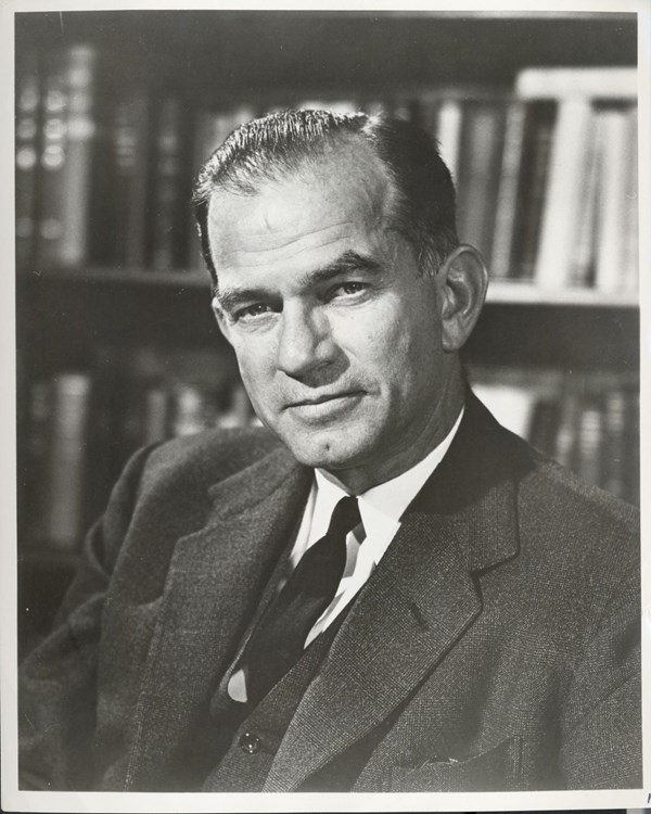 A black and white photograph of a man in a suit and tie sitting in front of a large bookshelf.