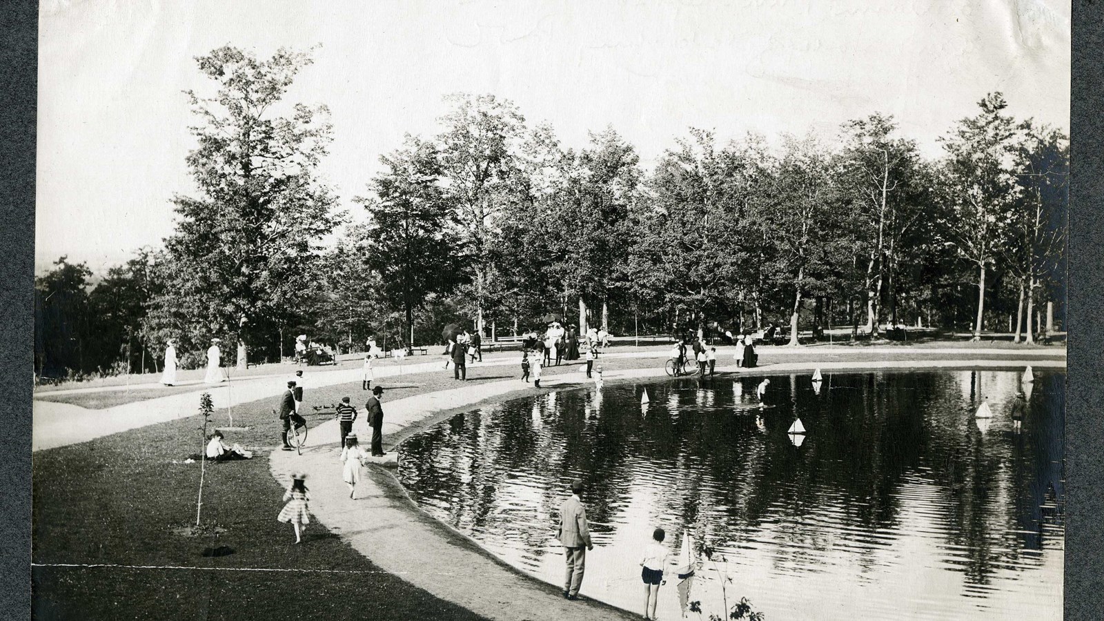 Black and white of water with people walking around, mini boats in it, paths and trees on edge