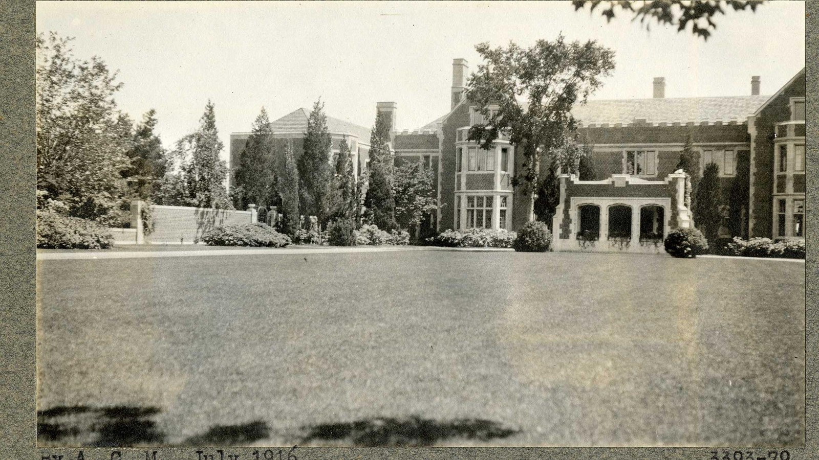 Black and white of flat grassy area in front of large brick house with trees and shrubs around it 