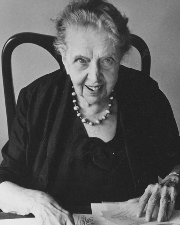 An elderly woman, grinning or speaking, looks up from a pile of papers