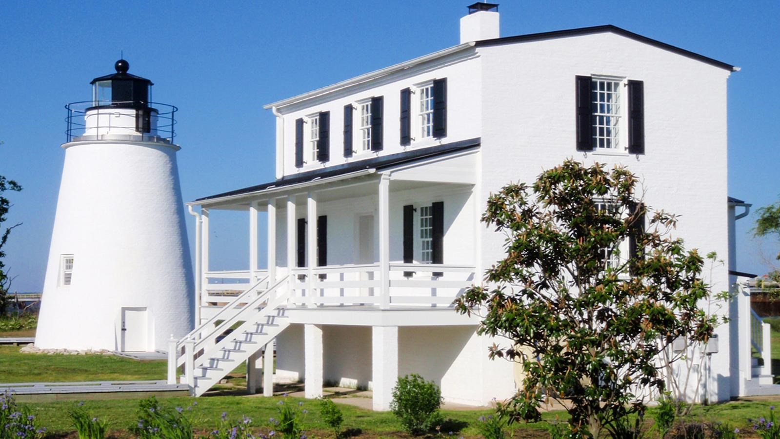 Piney Point Lighthouse and adjacent white brick building with gardens, under a clear blue sky.