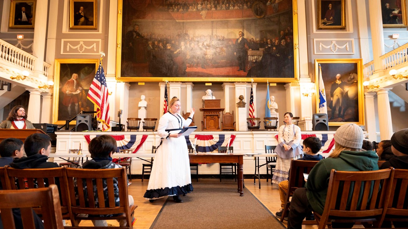 three adults in 1800s clothing stand in front of the stage Faneuil Hall as students look on.