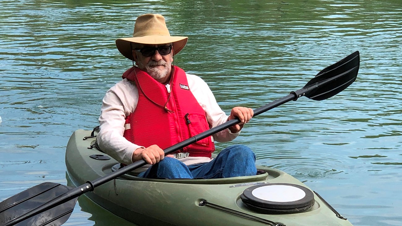 a man wearing hat, sunglasses and red pfd in a green kayak holding a black paddle on the water