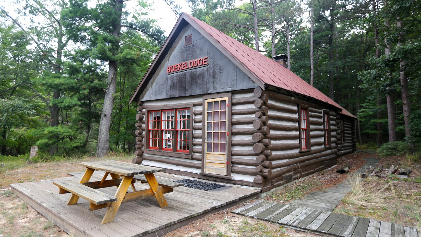 Log cabin with white mortar, a red roof, red window trim, a a wooden deck with a picnic table