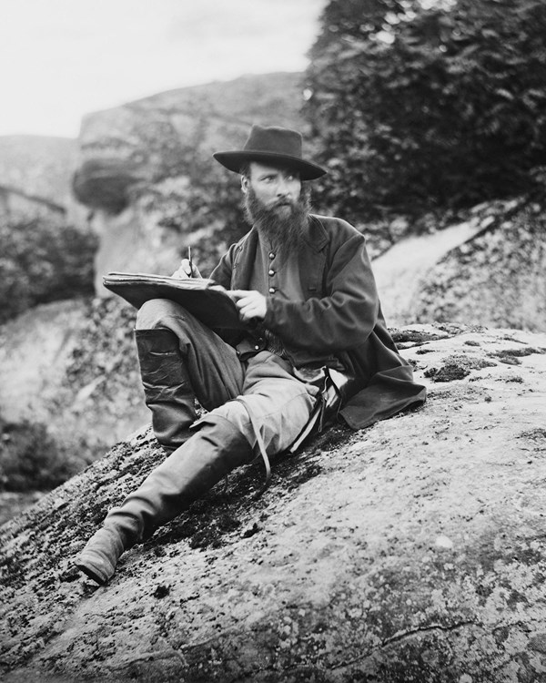 Black and white photo of man with sketch book. Man sits atop a boulder and looks to the side.