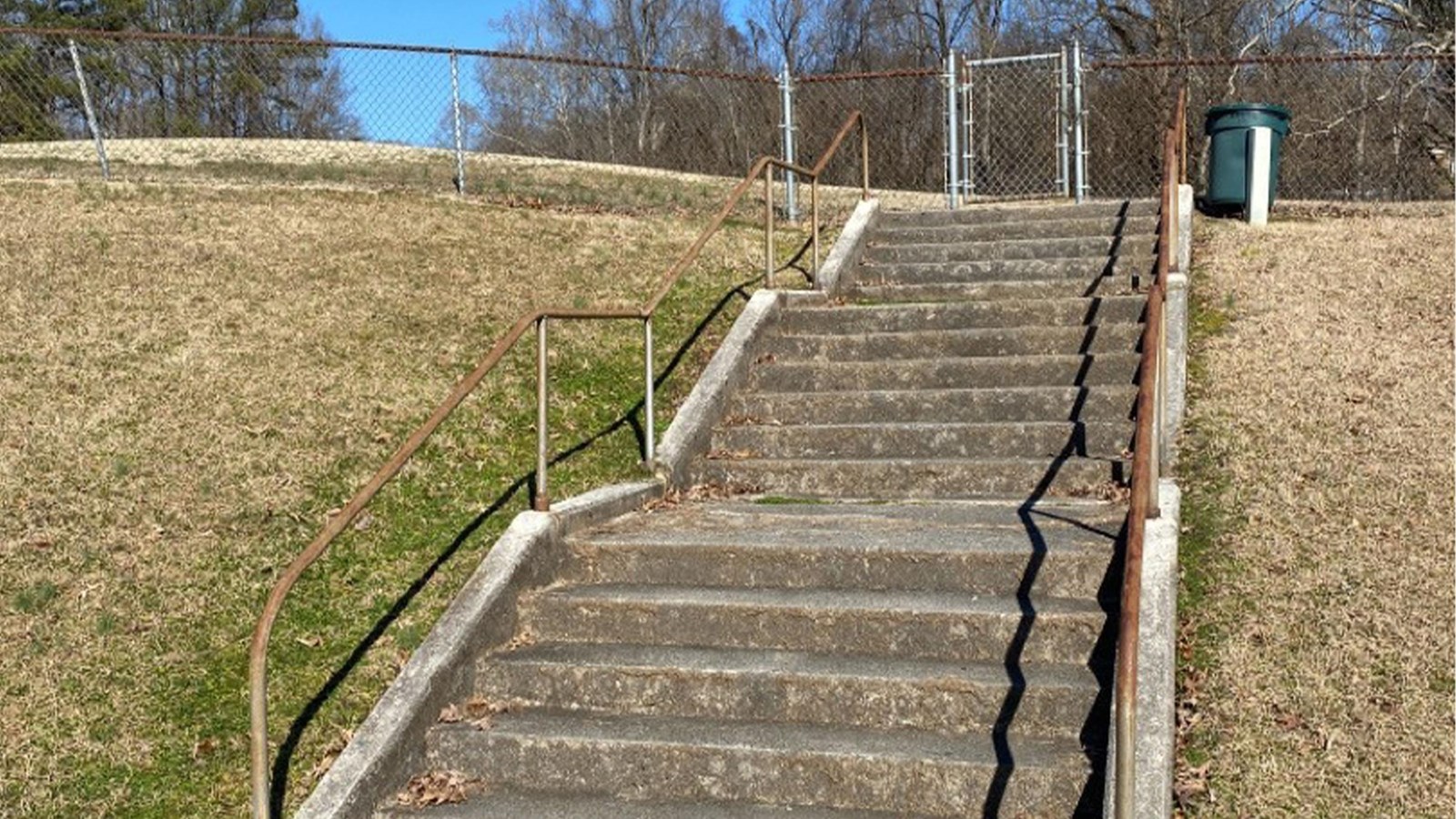 A concrete staircase with metal railing leads to a small grassy park with a chain link fence.