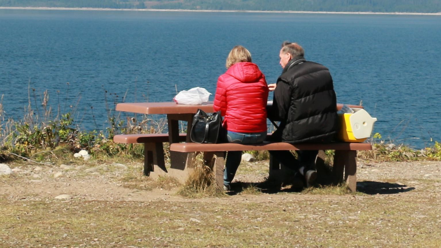 People sitting at picnic tables with a view of a blue lake and mountains in the background.
