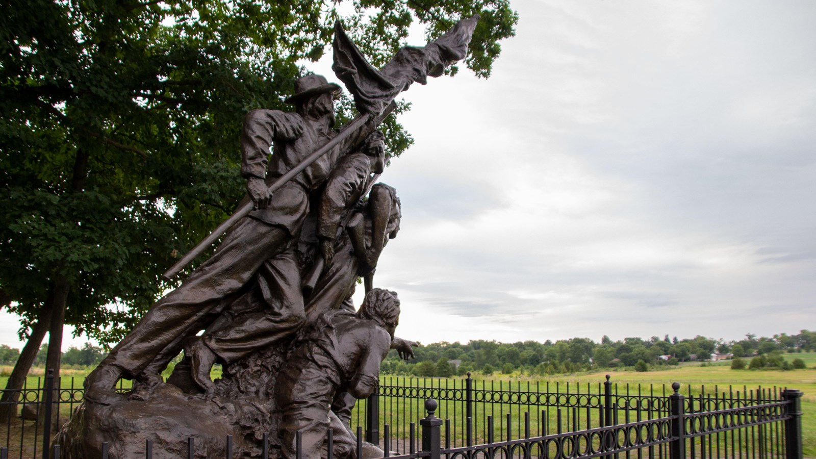 A bronze monument of soldiers charging forward towards a green farm field.